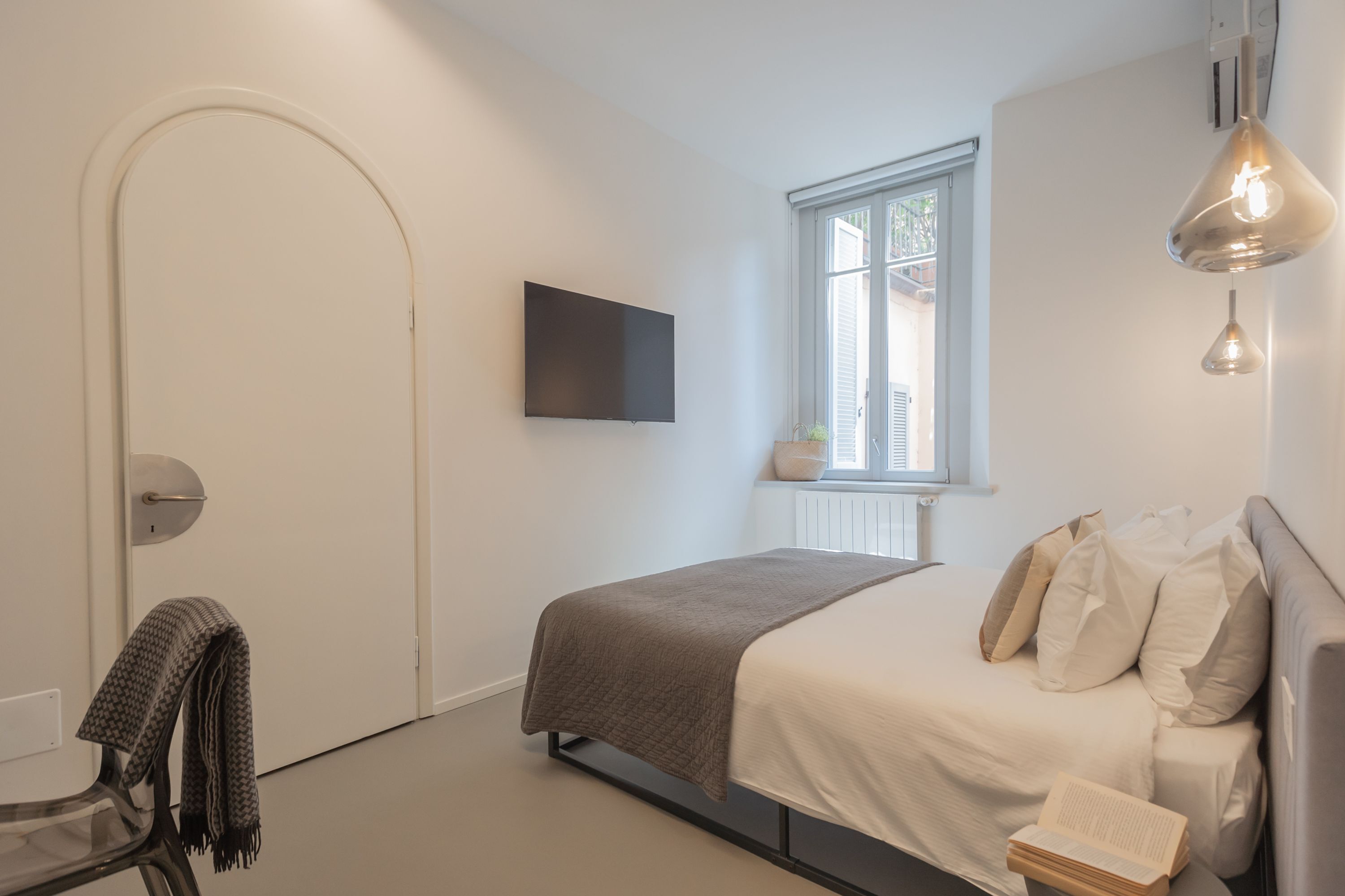 Un dormitorio moderno con cama, televisor, ventana y lámpara colgante.