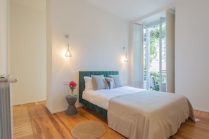 A bedroom with a teal headboard, a large bed, bedside lamps, a small round rug, and a window with a view of greenery. - Como Liberty Suites - Garbo (Como)
