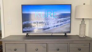 A television displaying a beach scene with the text "KURE BEACH" on a wooden dresser with drawers.