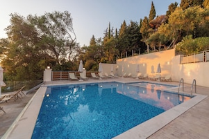 Una piscina rectangular con suelo de baldosas, rodeada de sillones de descanso y sombrillas, con árboles y un edificio al fondo.
