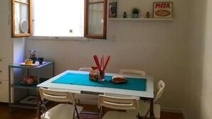 A dining area with a table set for two, a window with curtains, a shelf with small plants and a pizza sign, and a kitchen area with a shelf and a jar of nuts. - Neri 33 in Firenze With 3 Bedrooms and 2 Bathrooms (Florence)