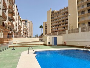 A swimming pool with a clear blue water, surrounded by a fence, and adjacent to a building with multiple balconies and a palm tree. - Alq 305 Calalga Apartment (Calpe)