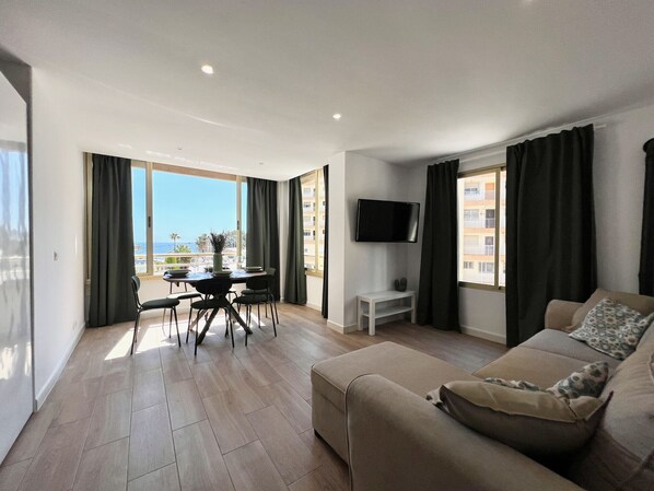 A modern living room with a sofa, a dining table with chairs, a TV mounted on the wall, and large windows with curtains. - Alq 305 Calalga Apartment (Calpe)