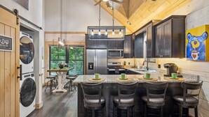 A modern kitchen with dark cabinets, a central island with bar stools, a ceiling fan, and a large window offering a view of greenery.