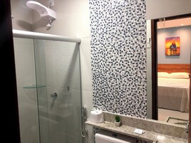 Un baño moderno con ducha de vidrio, pared con diseño, lavamanos con espejo y una cama visible a través del espejo.