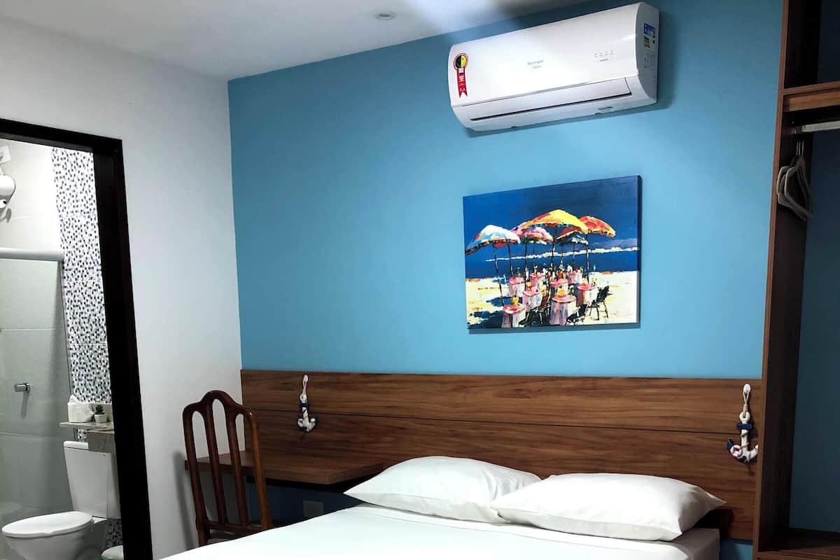 A hotel room with a bed, a wooden headboard, a painting of a beach scene, an air conditioner, and a small refrigerator.