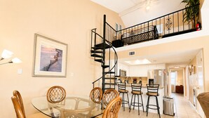 A dining area with a glass table and wicker chairs, a kitchen with white cabinets, a spiral staircase, and a framed picture on the wall.