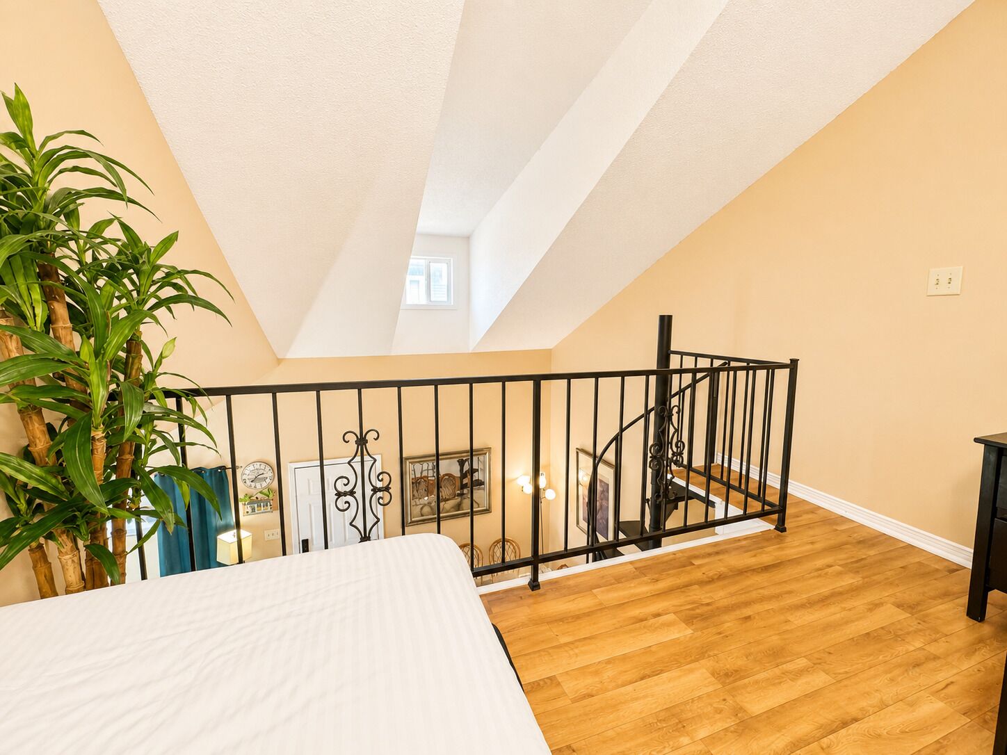 A bedroom with a wooden floor, a bed, a staircase with a black railing, and a potted plant.