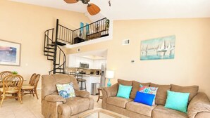 A living room with a sofa, two armchairs, a glass coffee table, a dining area with chairs, a kitchen, and a staircase leading to an upper level.
