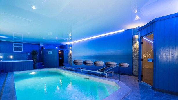 Una piscina interior con techo iluminado en azul, pared de piedra y sillas de descanso.