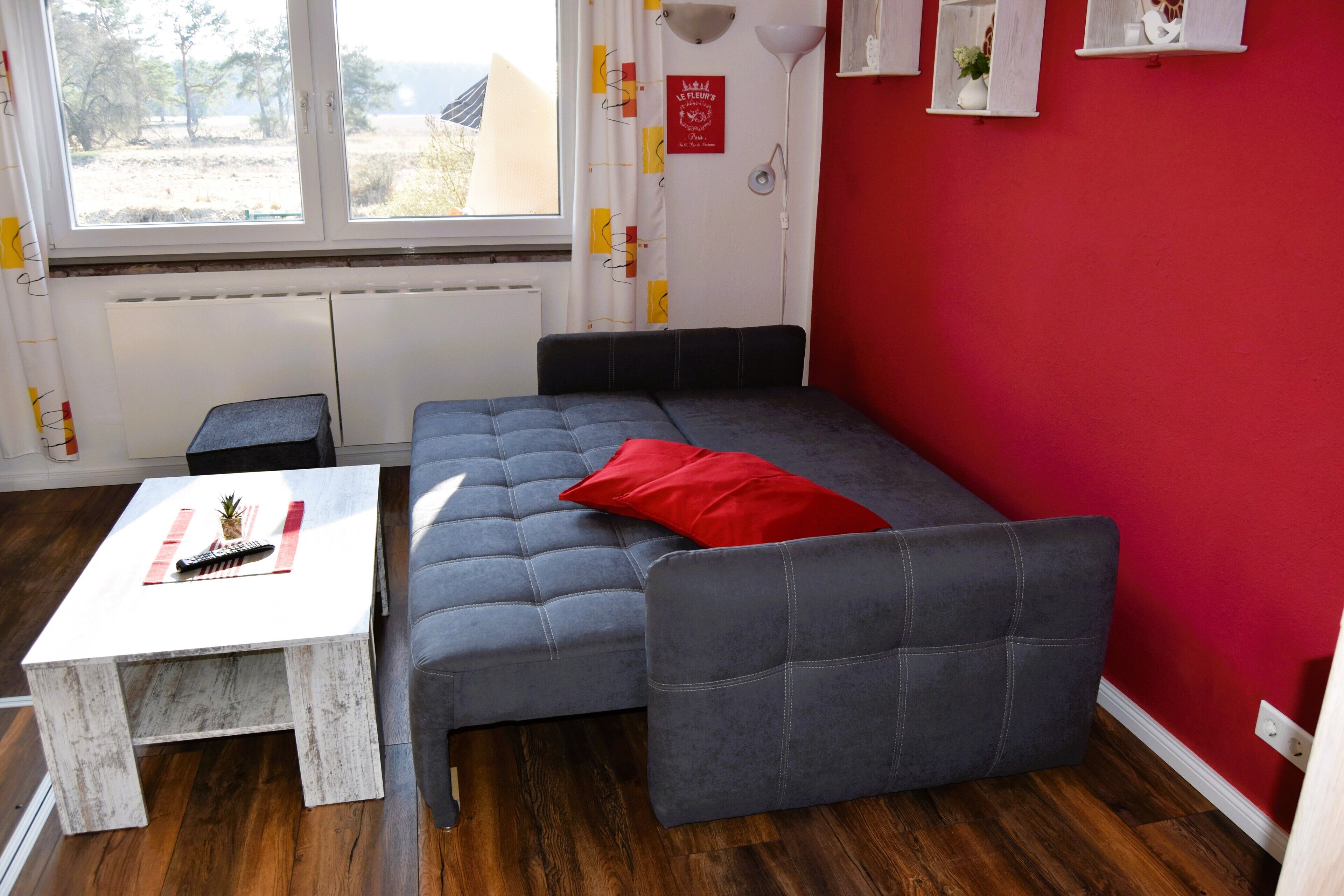 Une pièce compacte comprenant un canapé-lit, une petite table, une fenêtre avec vue et un mur rouge.