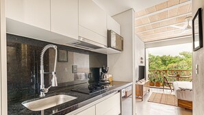 Cocina moderna con encimera negra, pileta de acero inoxidable y estufa integrada. Cuenta con un pequeño comedor con mesa y sillas, y un balcón con vista a la vegetación.