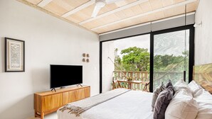 Un dormitorio con una cama grande, un televisor sobre un mueble de madera, un balcón con vistas a árboles y un cuadro enmarcado en la pared.