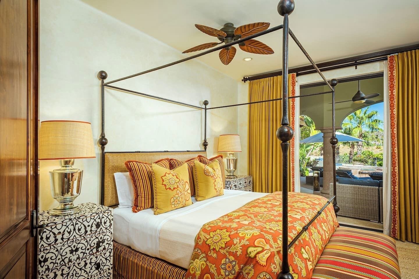 A four-poster bed with a ceiling fan, a balcony with a view of palm trees, and a patterned bedspread.