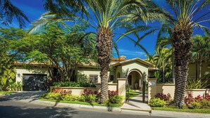 A residential property with a gated entrance, a driveway, and lush greenery including palm trees and various plants.