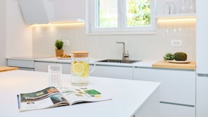 A modern kitchen with white cabinets, a window with a view of greenery, a kitchen island with a glass pitcher and a magazine, and a pineapple on the counter. - Hedera Estate, Villa Hedera Xviii (Zupa dubrovacka)
