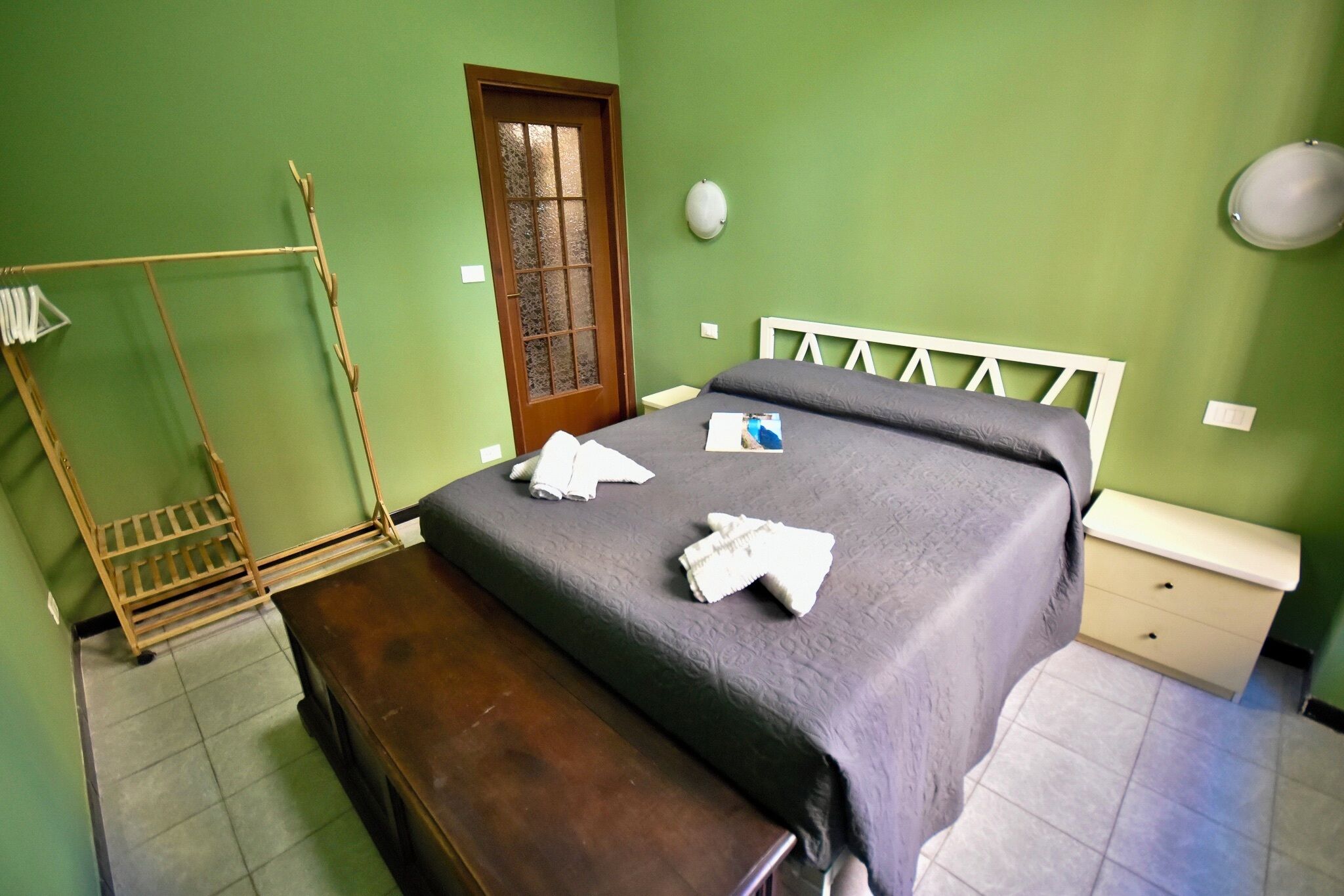 Una habitación con una cama, un banco de madera, un tendedero, una puerta y una mesita de noche.