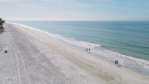 En strand med klar himmel, stilla hav och några få personer som promenerar längs strandkanten.