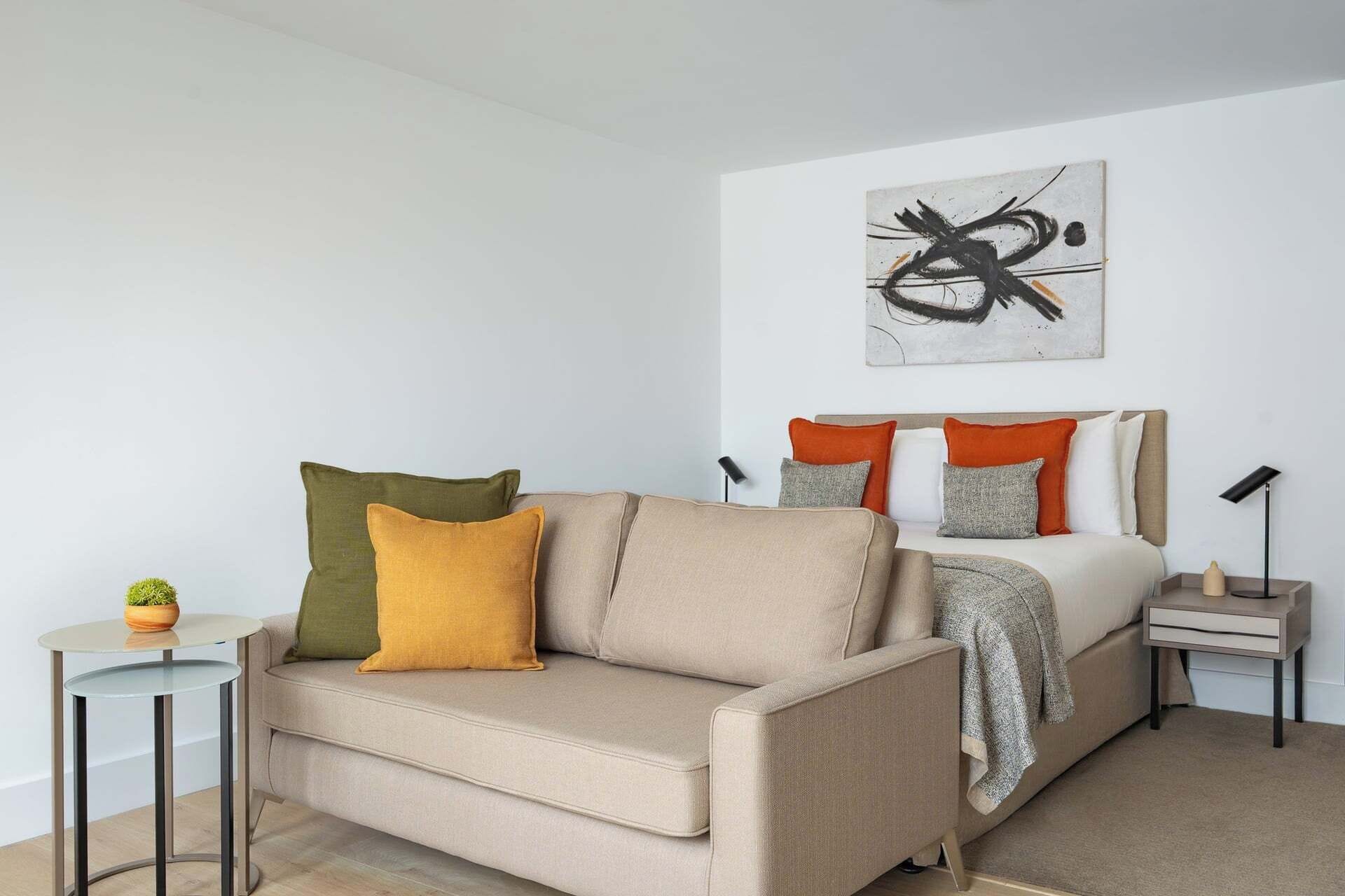A modern bedroom with a sofa, a bed with orange and grey pillows, a small table with a plant, and a wall art piece.