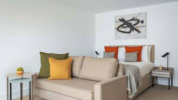 A modern bedroom with a sofa, a bed with orange and grey pillows, a small table with a plant, and a wall art piece.