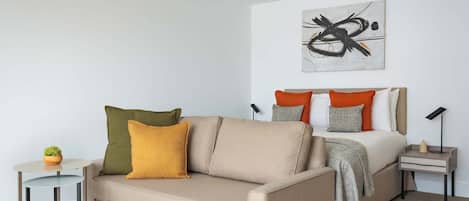 A modern bedroom with a sofa, a bed with orange and grey pillows, a small table with a plant, and a wall art piece.