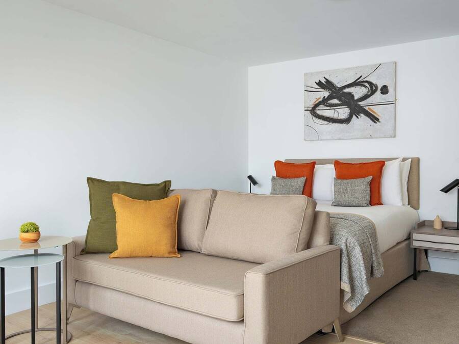 A modern bedroom with a sofa, a bed with orange and grey pillows, a small table with a plant, and a wall art piece.