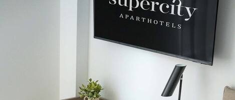 A modern hotel room with a bed, a desk with a laptop, a chair, a small plant, and a wall-mounted TV displaying 'Supercity Apart Hotels'.