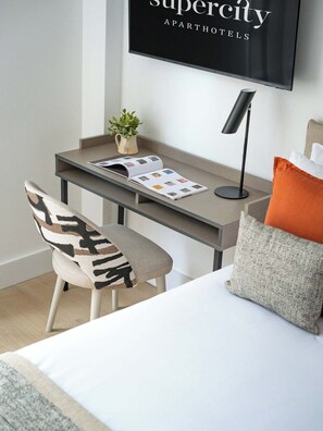 Una habitación de hotel moderna con una cama, un escritorio con una computadora portátil, una silla, una planta pequeña y un televisor de pared que muestra 'Supercity Apart Hotels'.