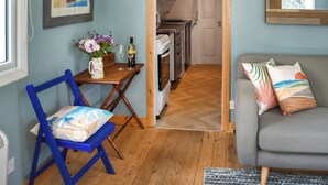 A living room with a sofa, a chair, a table with a vase of flowers, and a kitchen visible through an open doorway. - Seaglass Chalet - UK47636 (Mundesley, near North Walsham)