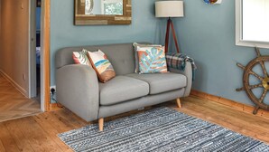 A living room with a grey sofa, patterned cushions, a striped rug, a wooden floor, a wall clock, a lamp, and a ship's wheel decoration. - Seaglass Chalet - UK47636 (Mundesley, near North Walsham)