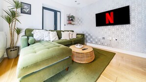Un salon moderne avec un canapé vert, un pouf rond en bois, un tapis vert et un téléviseur fixé au mur qui affiche le logo de Netflix.