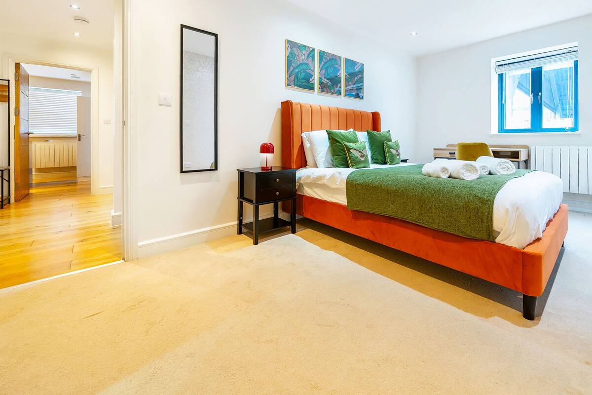 A modern bedroom with a wooden floor, a bed with orange headboard, green and white bedding, a nightstand, and a window with blinds.