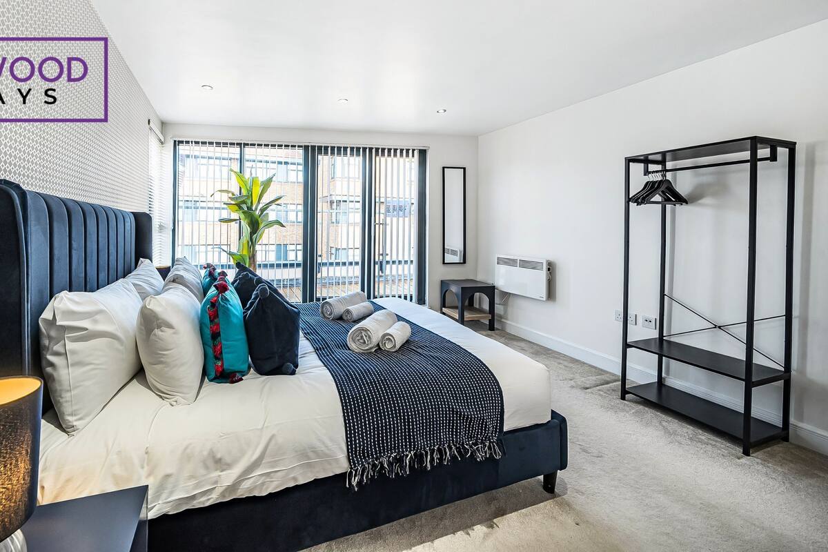 A modern bedroom with a large bed, a side table, a hanging wardrobe, and a view of the city through large windows.