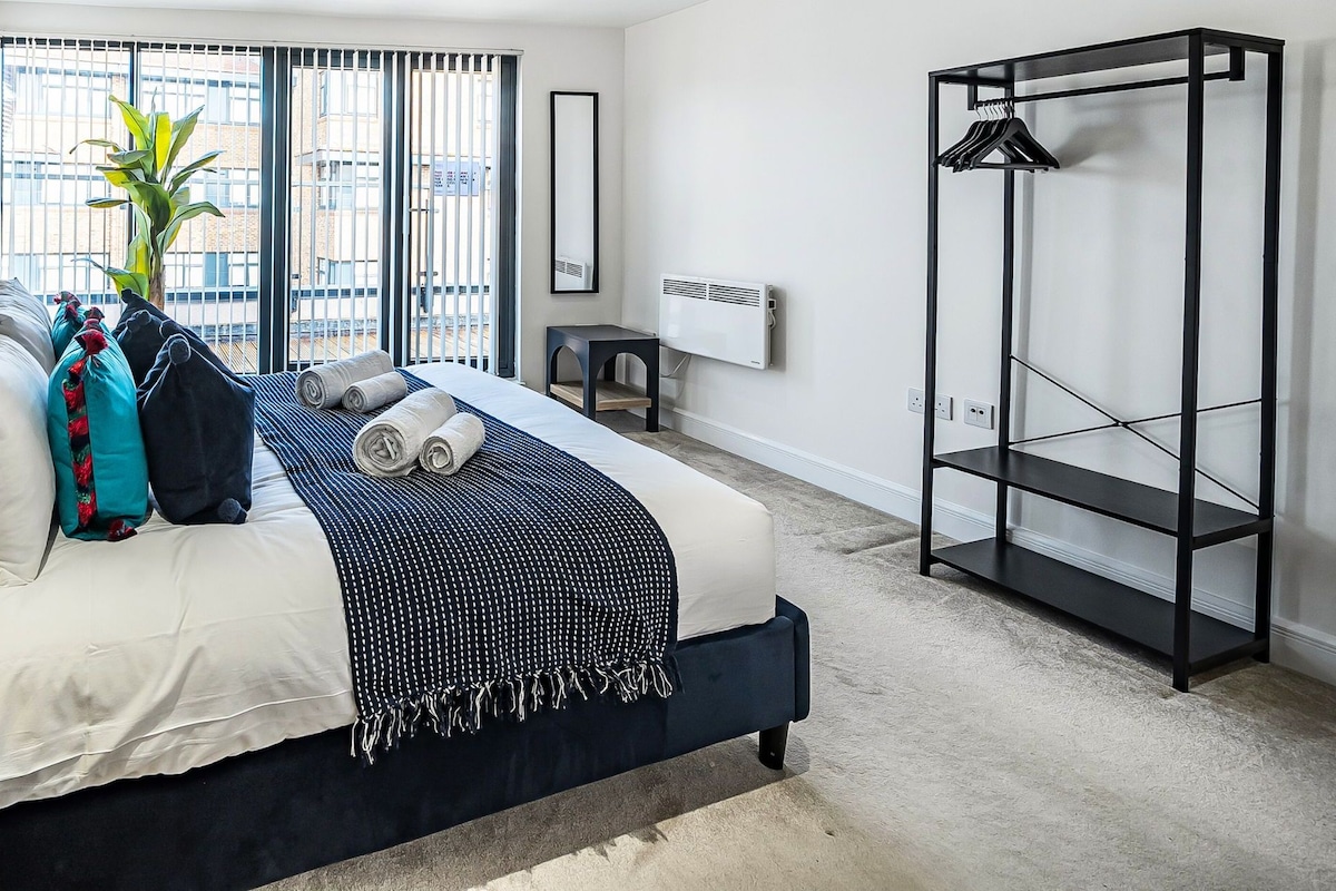 A modern bedroom with a large bed, a side table, a hanging wardrobe, and a view of the city through large windows.