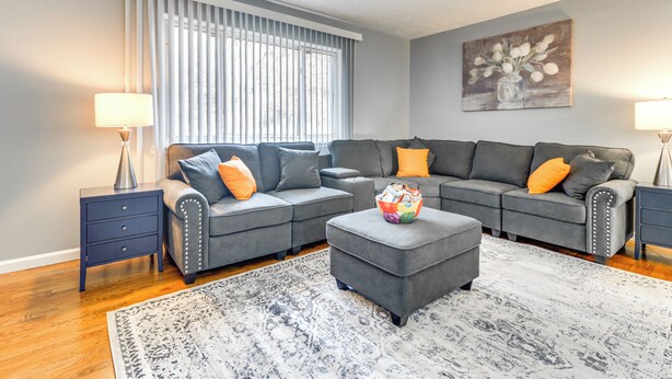 Ein Wohnzimmer mit einem grauen Ecksofa, einem passenden Hocker, einem gemusterten Teppich und einem Bild an der Wand.