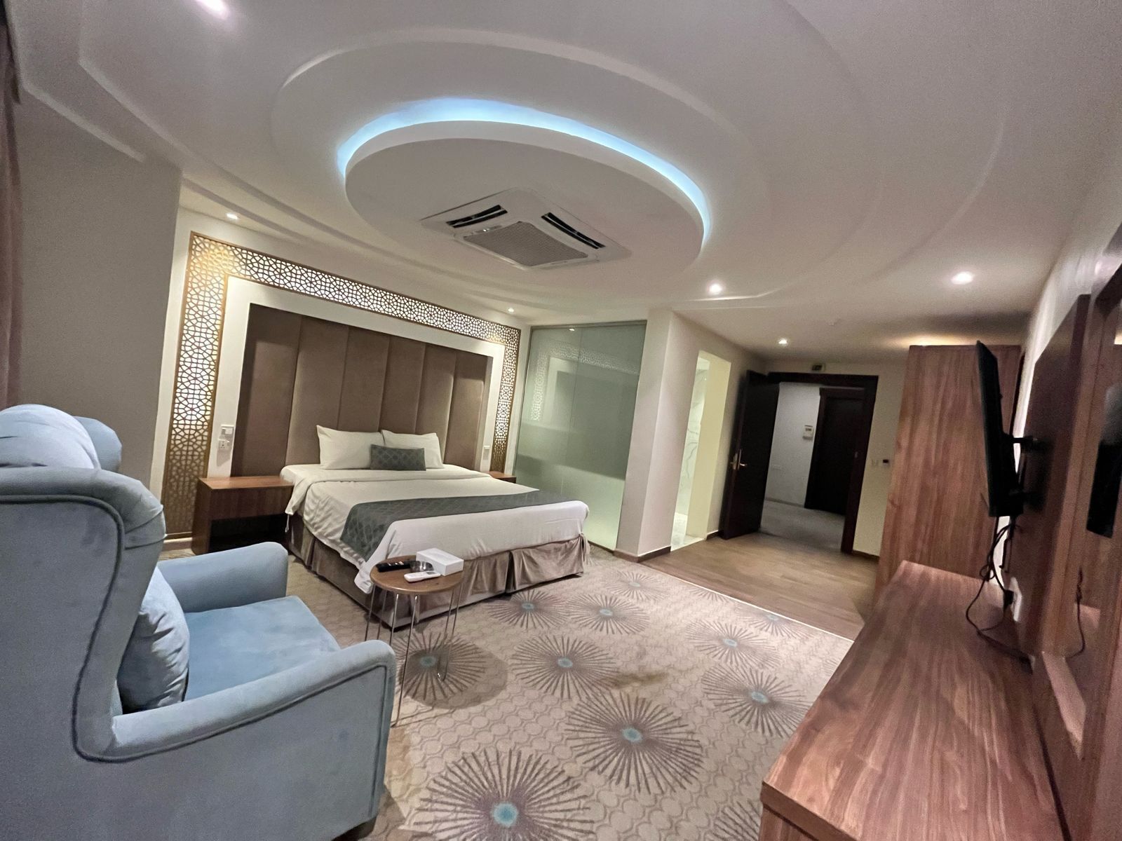 A modern hotel room with a large bed, a seating area with a sofa and armchair, a wooden side table, and a flat-screen TV mounted on the wall.