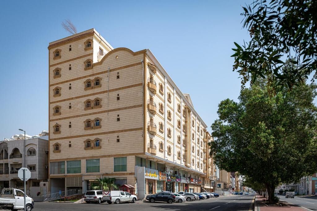 A multi-story hotel with a beige facade, multiple balconies, and a row of shops on the ground floor.