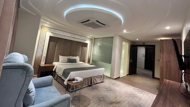 A modern hotel room with a large bed, a seating area with a chair, a small table, and a decorative wall panel. - Al Salamh suites Family - أجنحة السلامة العائلية (Jeddah)