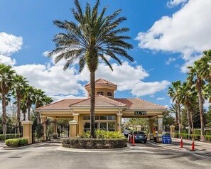 Entrada de hotel com palmeira, portão e um carro estacionado.