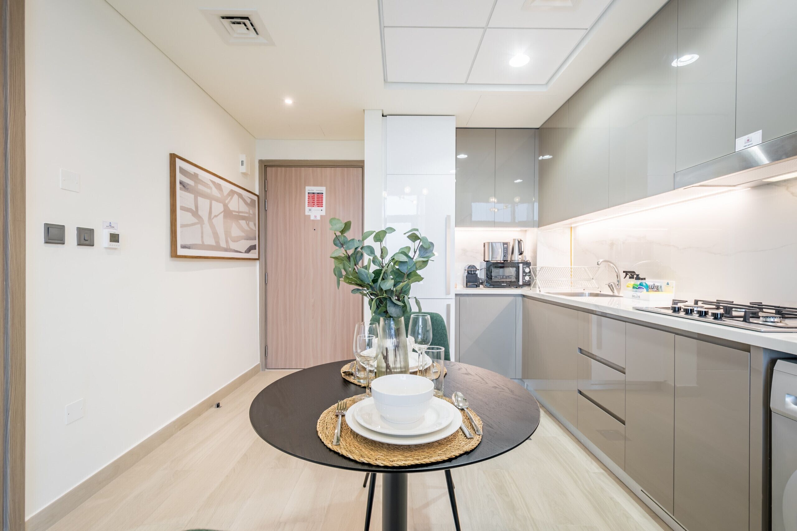 A modern kitchen with a dining area, including a round table with a white bowl, plates, and cutlery, a vase with green plants, and a microwave.