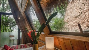 Una habitación con techo de madera, una ventana con vista a follaje, una mesita de noche con lámpara y una planta.