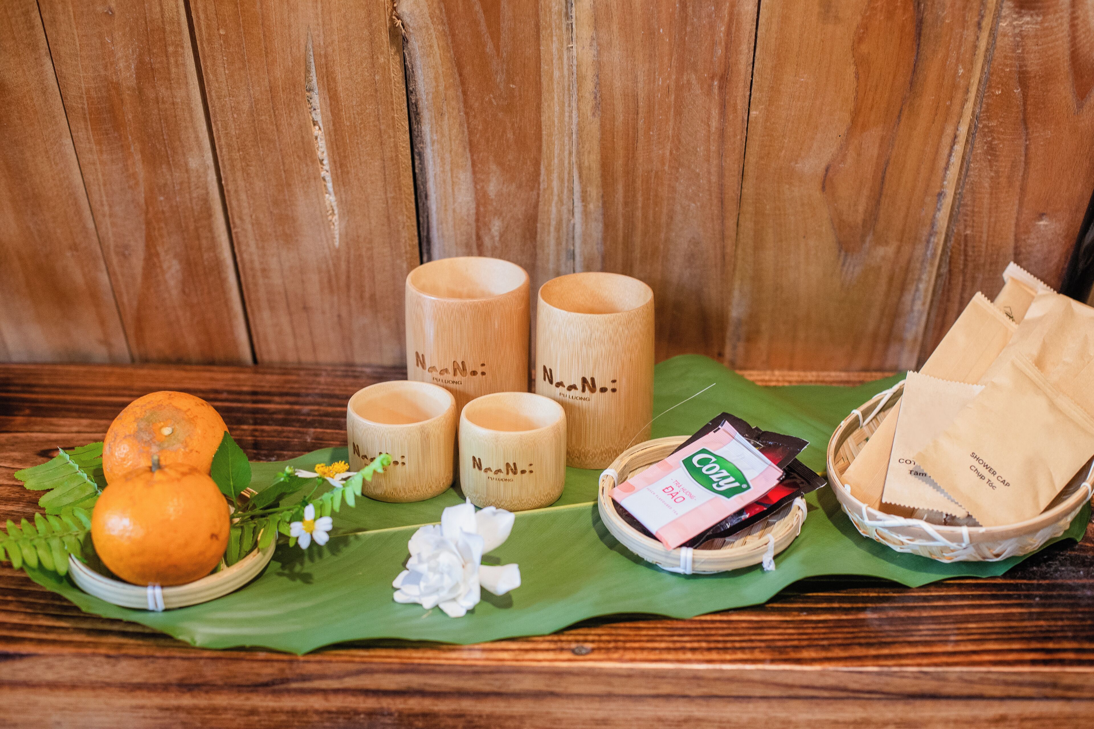 A wooden table with a green leaf, two oranges, three wooden cups, a basket with paper bags, and a small packet.