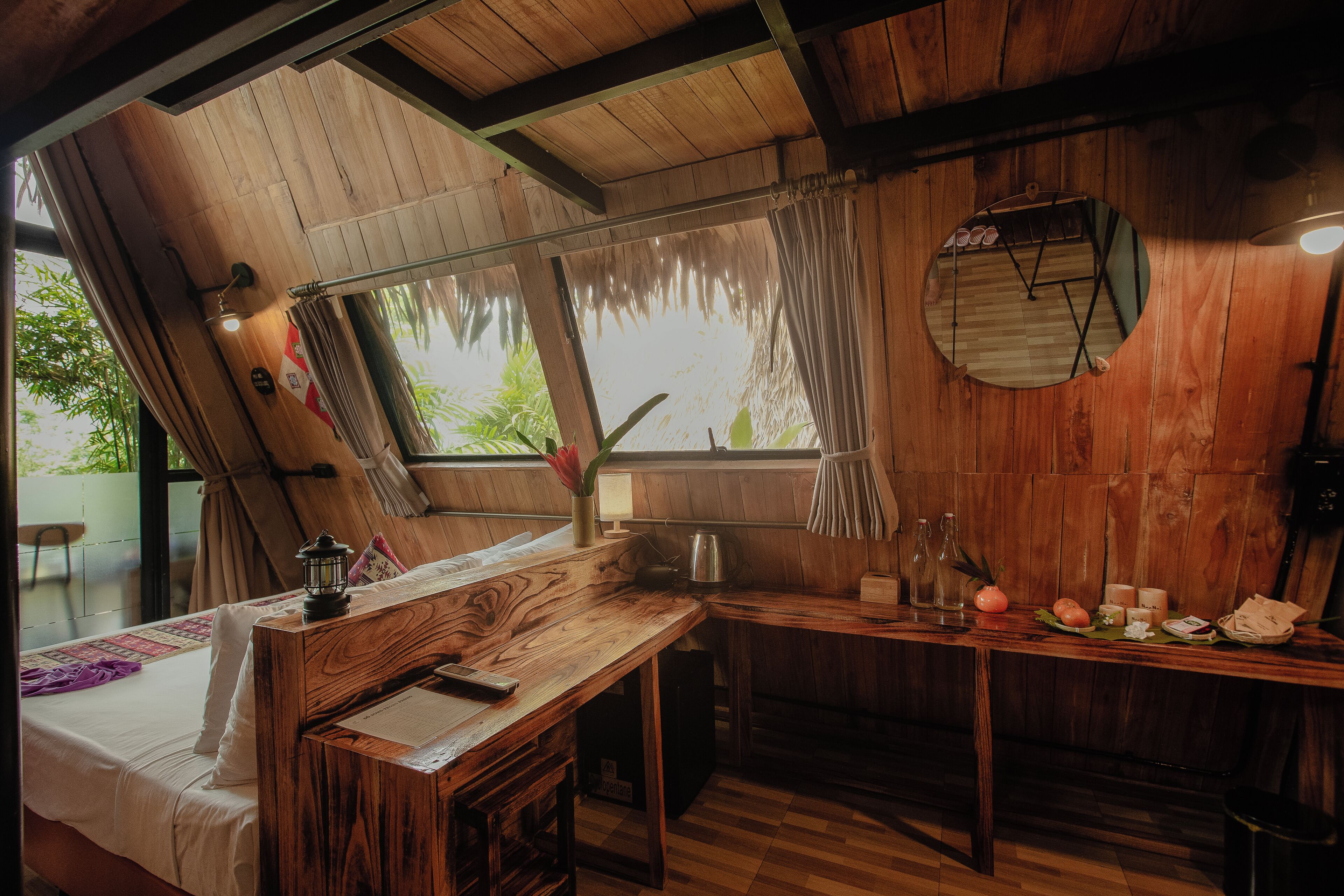 A wooden room with a bed, a desk, a mirror, and a window with curtains.
