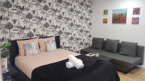 Une chambre avec un lit, un canapé, un mur recouvert d’un papier peint à motifs tropicaux et une petite table sur laquelle est posée une plante.