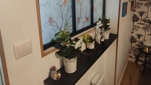 Une fenêtre ornée d’un motif coloré représentant des oiseaux, une étagère noire avec des plantes et un mur blanc décoré d’un motif de palmiers.