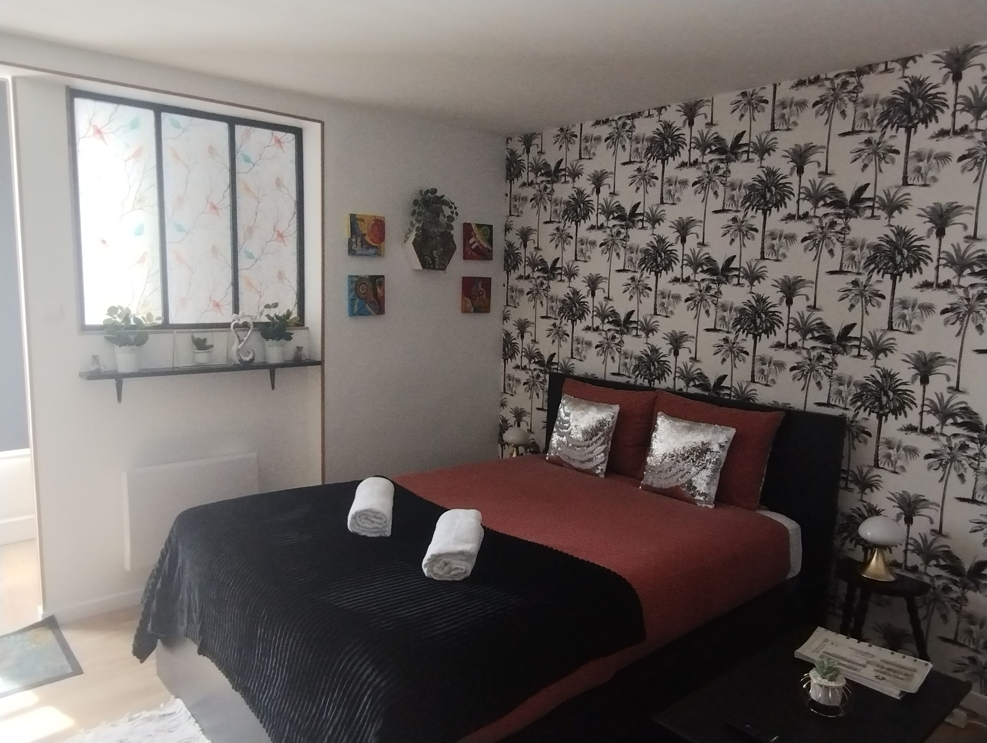 A bedroom with a bed, a window with a view, a wall with a palm tree pattern, and a small table with a lamp.