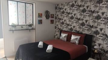 A bedroom with a bed, a window with a view, a wall with a palm tree pattern, and a small table with a lamp.