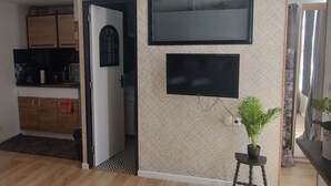 Un salon moderne avec un téléviseur à écran plat fixé au mur, une plante en pot posée sur une petite table et un coin cuisine visible à travers une porte ouverte.