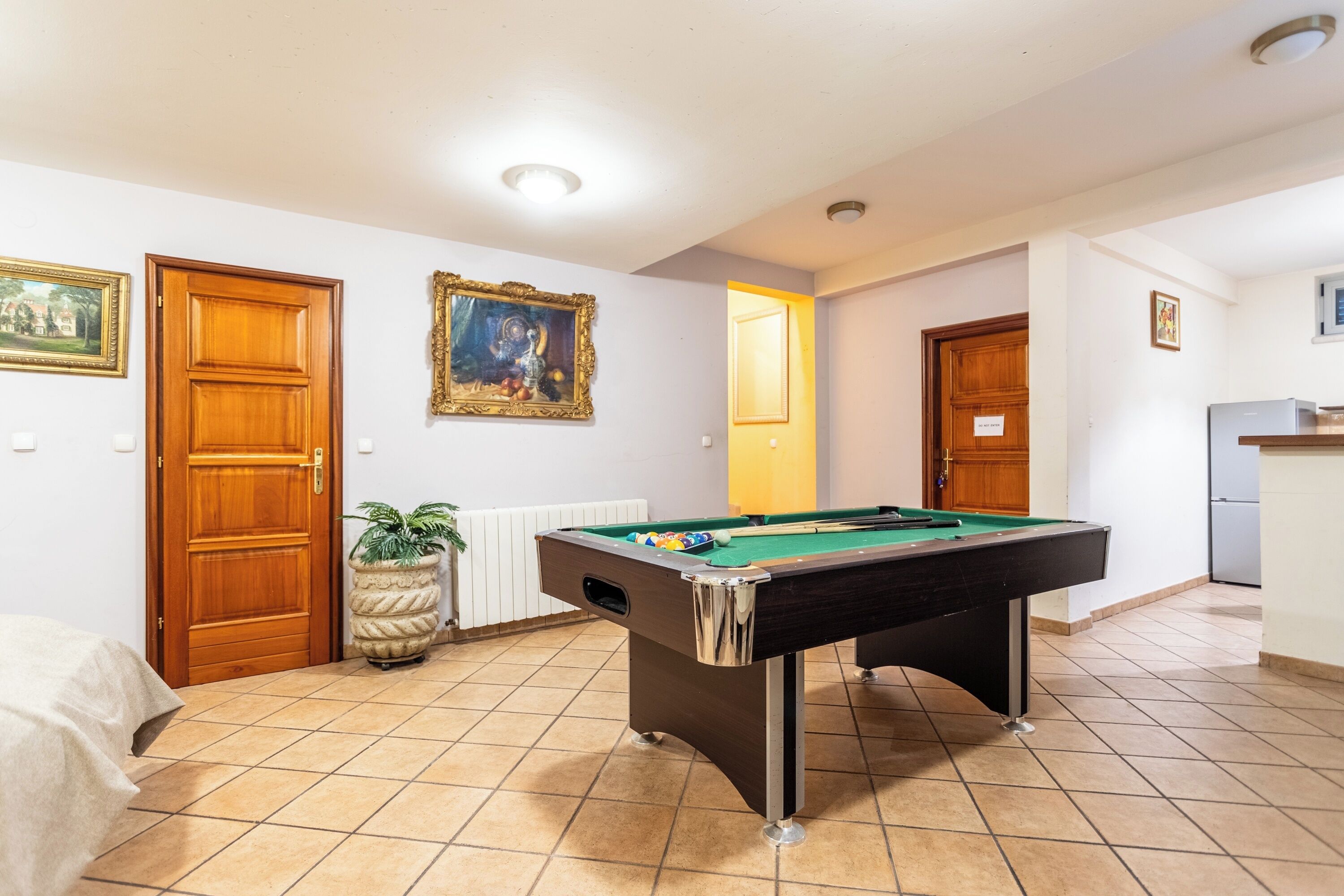 A pool table in a room with tiled flooring, wooden doors, and a small kitchen area.