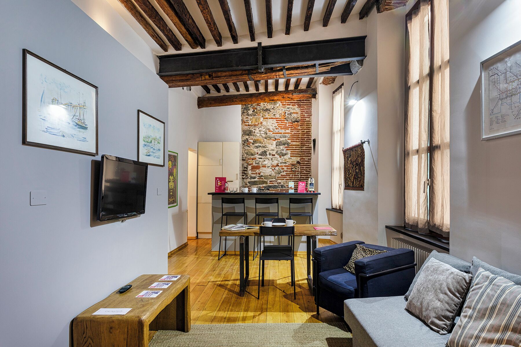 A cozy living area with a wooden bench, a sofa, and a dining table with chairs. A kitchen area is visible in the background with a brick wall and a window.